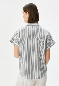 Camicetta a maniche corte con colletto in tessuto leggero, caratterizzata da strisce verticali grigie, blu navy e rosa, con orlo leggermente curvo e taschino sul retro.
