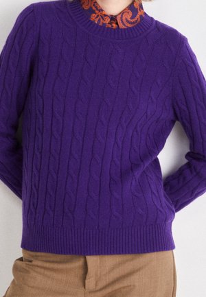 Violetti palmikkoneule, jossa on ribbaus kauluksessa ja helmassa, päällekkäin kuvioidun oranssin kauluspuseron kanssa, joka on sileätekstuurinen.