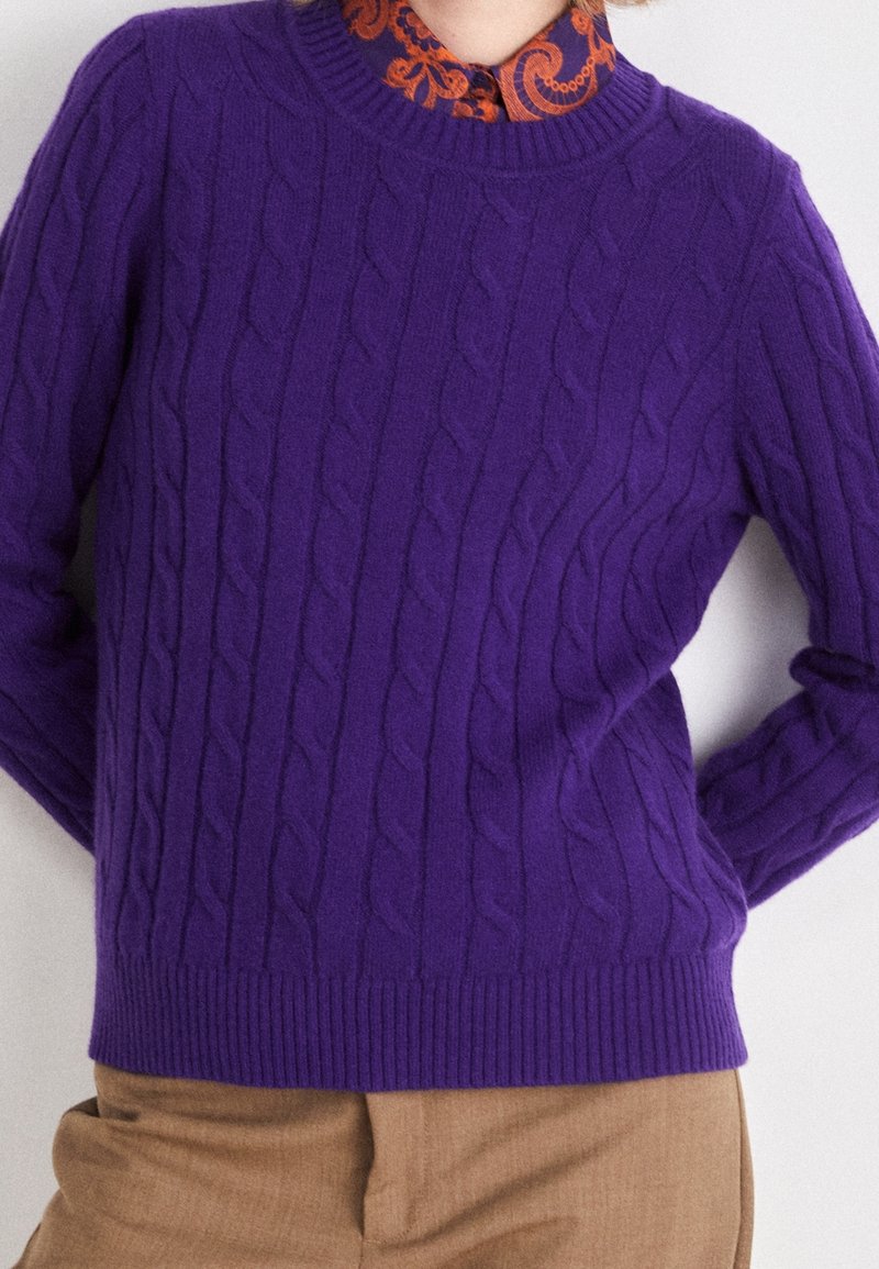 Lila kabelstickad tröja med en ribbad halsringning och fåll, lagd över en mönstrad orange skjorta med krage, som har en slät textur.