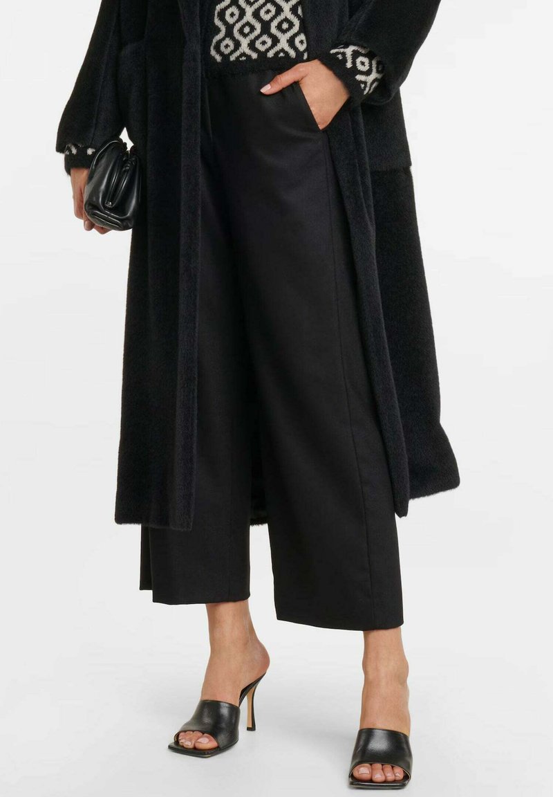 Zwarte wollen coat met geometrisch patroon stof, brede zwarte broek en zwarte hakken mules. Opvallende details zijn de zachte textuur en het slanke design.