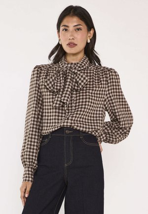 Donna che indossa una camicetta marrone a quadri con un grande fiocco al collo e jeans blu scuro a vita alta su uno sfondo semplice.