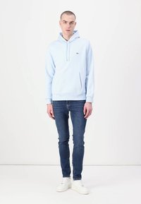 Sudadera de color azul claro con cordón, bolsillo frontal y logo; combinada con unos jeans ajustados de color azul oscuro y zapatillas blancas, de pie contra un fondo blanco.