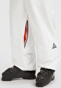Pantalones de esquí blancos con cremalleras que revelan un forro de malla naranja, combinados con botas de esquí negras con acentos metálicos y cierres ajustables.