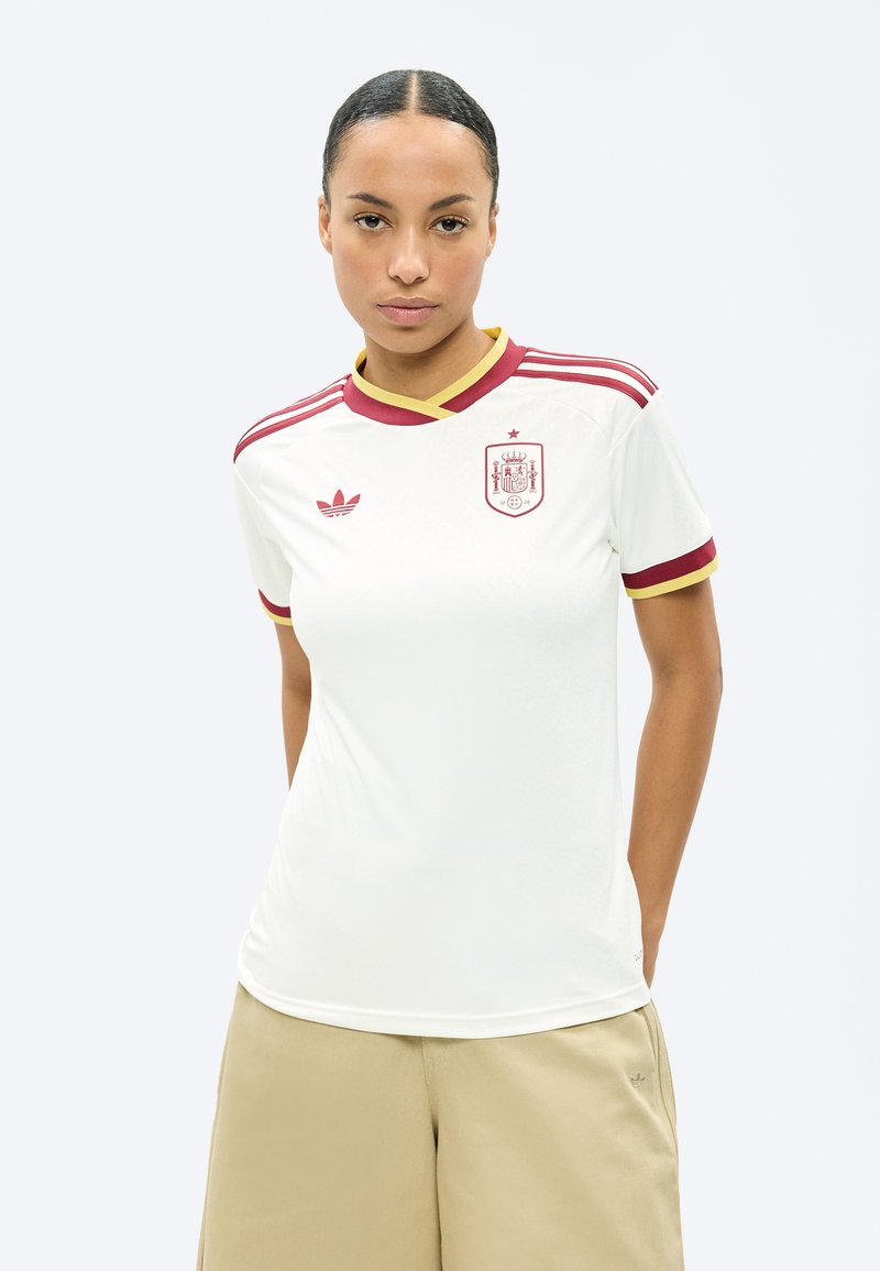 Γυναίκα που φοράει λευκή φανέλα ποδοσφαίρου της Ισπανίας με κόκκινες και κίτρινες αποχρώσεις, στέκεται με τα χέρια πίσω από την πλάτη.