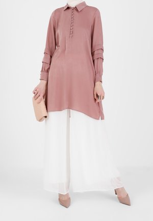 Femme portant une tunique rose poudré à manches longues avec des détails de boutons, un pantalon blanc large, des talons couleur peau et tenant une pochette beige.