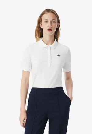 Polo bianco con placca di bottoni e maniche corte, con logo di coccodrillo verde sul lato sinistro del petto. Indossato con pantaloni blu navy.