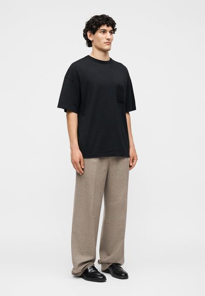 Άνδρας που στέκεται φορώντας μαύρο υπερμεγέθη μπλουζάκι με υφασμένη τσέπη, μπεζ παντελόνι με φαρδιά πόδια και μαύρα δερμάτινα παπούτσια πάνω σε λευκό φόντο.