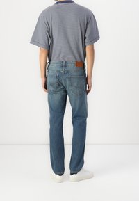 Camisa polo de rayas para hombre en azul marino y blanco, combinada con jeans de mezclilla azul que tienen un parche de cuero y dos bolsillos traseros, llevada con zapatillas blancas.