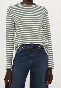 Zeleno-bíle pruhovaná košile s dlouhým rukávem vyrobená z měkké látky, v kombinaci s tmavě modrými džíny s knoflíkovým zapínáním a jemným prošíváním.