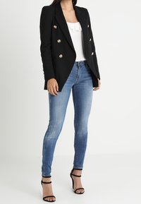 Black double-breasted blazer with gold buttons, white ruffled top, and blue skinny jeans. The model wears black strappy high heels.
