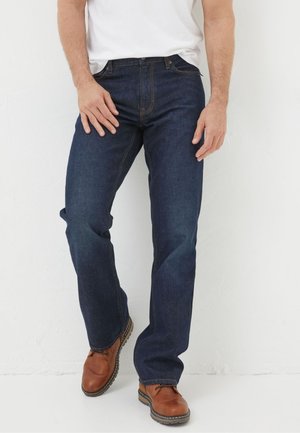 Mann, der dunkelblaue, gerade geschnittene Jeans, weißes T-Shirt und braune Lederschuhe trägt, steht vor weißem Hintergrund.