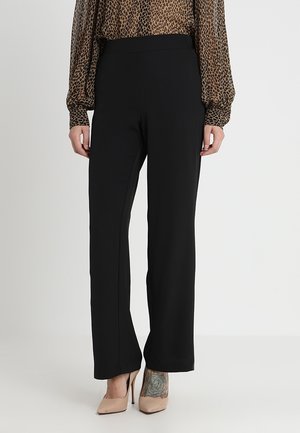 Femme portant un pantalon noir à jambes droites, des talons hauts beige et un chemisier à manches longues marron à imprimé léopard, debout devant un fond uni.