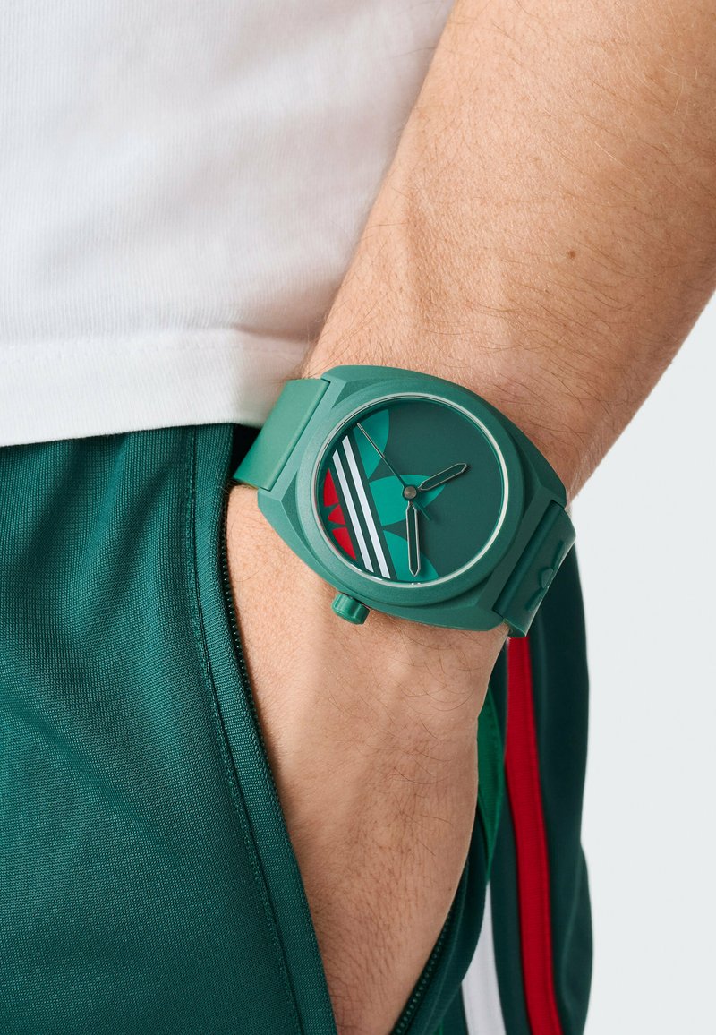 Grøn silikoneur med rund urskive, som har en ensfarvet grøn skive, minimalistiske timeindikatorer og røde og hvide designelementer.