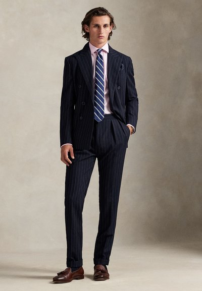 Tamsiai mėlynas dvieilis kostiumas su juostelėmis, pritaikytas figūrai, su šviesiai rožine palaidine, mėlynos spalvos dryžuota kaklaraište ir rudos odos loaferiais, su kišeniniu rankšluosčiu.