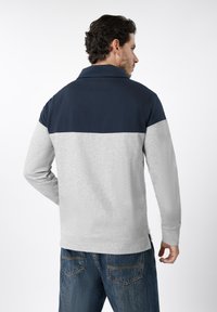 Άνδρας που φοράει μακρυμάνικη μπλούζα δύο τόνων σε ναυτικό μπλε και ανοιχτό γκρι με μπλε τζιν, γυρισμένος με την πλάτη προς τα πίσω, πάνω σε απλό λευκό φόντο.