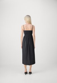 Robe noire longueur genoux avec un corsage smocké et des fines bretelles, présentant une silhouette ample. Portée avec des chaussures à talons hauts noires.
