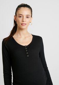 Top preto canelado de mangas longas com decote em V, apresentando cinco botões como destaque na parte da frente. Textura suave e design ajustado.