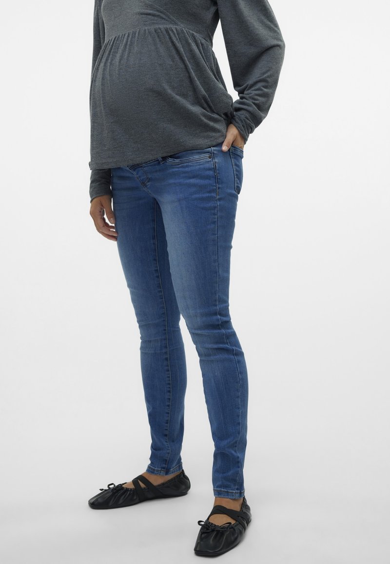 Jeans skinny bleu de maternité, en tissu denim, design ajusté, taille mi-haute, associés à des chaussures plates noires avec un détail de nœud.