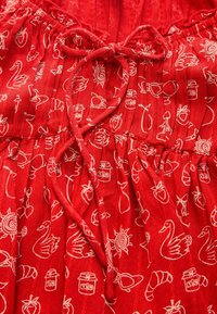 Červená látka s bílými ilustracemi různých předmětů, včetně labutí, ovoce a dezertů. Obsahuje nařasenou elastickou část a zavazovací detail.