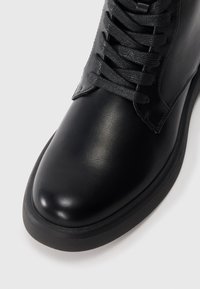 Botín negro de cuero hasta el tobillo con un acabado suave, puntera redonda y cordones texturizados. Presenta una suela de goma y acentos de costura mínimos.