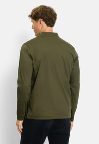 Langarm-Polohemd in Olivgrün, aus weichem Stoff. Verfügt über einen klassischen Kragen, seitliche Schlitze und enganliegende Manschetten. Einfaches Design mit minimalen Details.