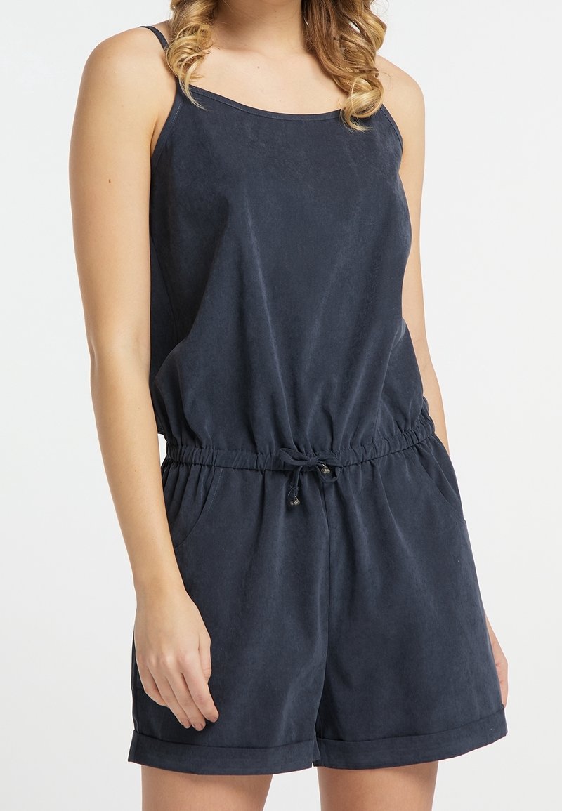 Femme portant une combinaison sans manches bleu foncé avec taille réglable par cordon et poches, debout devant un fond blanc uni.