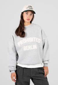 Fiatal nő világosszürke pulóverben, rajta a "PROHIBITED BERLIN" felirattal és megfelelő sapkával, sötétszürke nadrággal és arany gyűrűkkel párosítva.