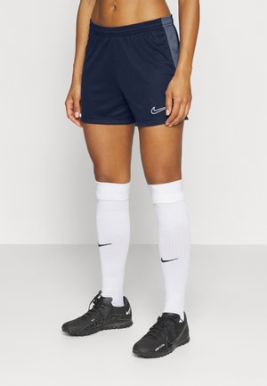 Person iført mørkeblå Nike sportsshorts, hvide knæhøje Nike sokker og sorte Nike sportssko står på ensfarvet baggrund.