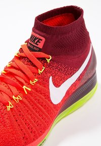 Červená a oranžová pletená sportovní bota s vysokým límcem, bílým logem Nike a žlutými tkaničkami. Má texturovaný svršek a zelenou podrážku.