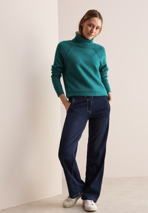 Maglione turtleneck turchese con dettagli a coste, abbinato a jeans a gamba larga blu scuro e sneaker bianche, in piedi davanti a uno sfondo neutro.