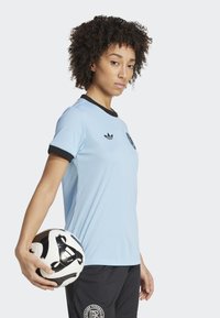 Lys blå fotballtrøye med svart krage og mansjetter, strukturert stoff, logo på brystet, brukt med mørke shorts, holder en fotball.