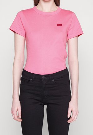 Kobieta ubrana w dopasowany różowy T-shirt z krótkim rękawem z małym czerwonym logo na piersi, połączony z czarnymi dżinsami z wysokim stanem.