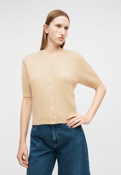 Γυναίκα με ίσια μαλλιά φορώντας μπεζ κοντομάνικη ζακέτα με κουμπιά και μπλε τζιν ψηλόμεσο, ποζάρει με το ένα χέρι στη μέση.