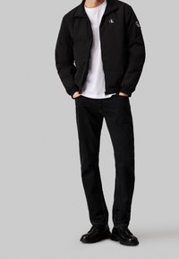 Man klädd i svart zip-up jacka med "cK" logotyper, vit t-shirt, svarta jeans och svarta läderskor, som står med händerna i fickorna.