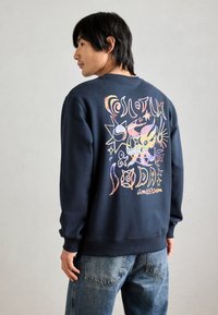 Sweatshirt marine avec un design graphique coloré comportant des étoiles et des formes abstraites au dos. Fabriqué en tissu en coton doux. Poignets et ourlet côtelés.