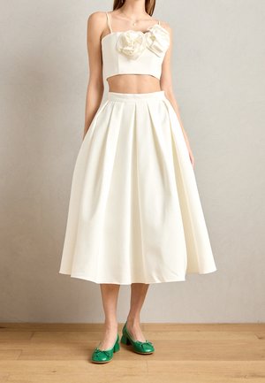 Mujer con top corto blanco sin tirantes con dos aplicaciones florales, falda midi plisada de talle alto y zapatos verdes de tacón grueso sobre suelo de madera.