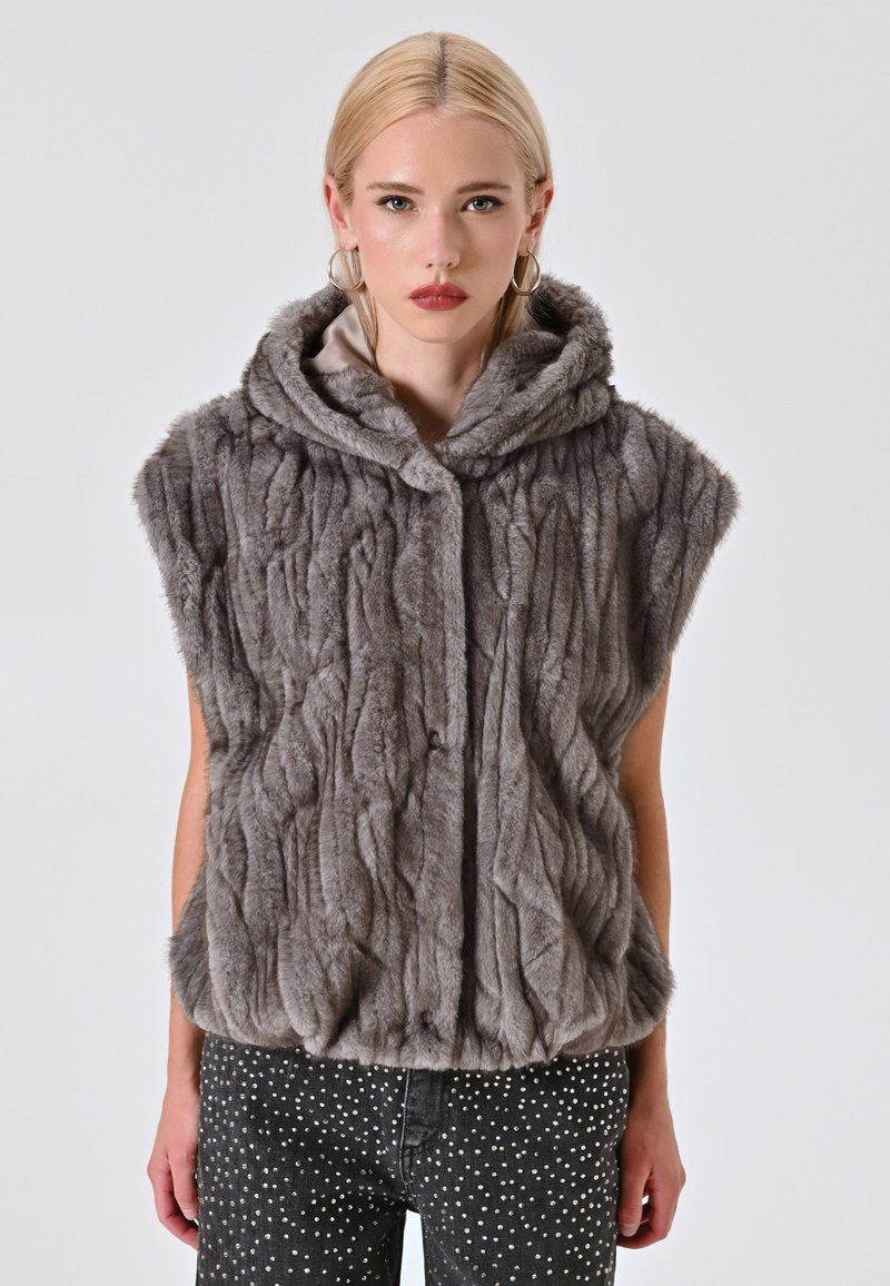Gilet in pelliccia sintetica grigia, con cappuccio, motivo testurizzato e chiusure frontali. Indossato con jeans neri decorati con pois argentati.