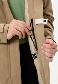 Mani che tengono aperto un giubbotto beige con una tasca nera con cerniera all'interno, mostrando un'etichetta in tessuto blu Novalon e una fascetta bianca con bottone.