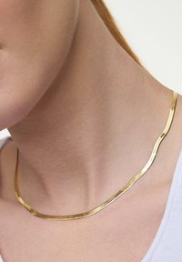 Złoty naszyjnik o falistym, płaskim designie, z teksturowaną powierzchnią. Końce delikatnie się zwężają i opierają blisko obojczyka.