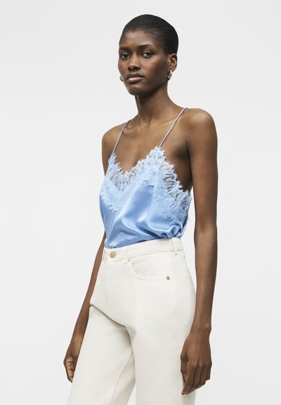 Γυναίκα που φοράει ανοιχτό μπλε σατέν καμιζόλη με δαντελένιο τελείωμα και λεπτές τιράντες, η οποία είναι μαζεμένη μέσα σε ψηλόμεσο εκρού παντελόνι, σε απλό φόντο.