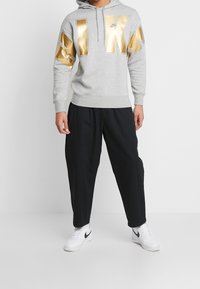 Grå hoodie med stora metalliska guld "NIKE" bokstäver, känguruficka och justerbar huva; tillsammans med svarta baggy-byxor och vita sneakers.