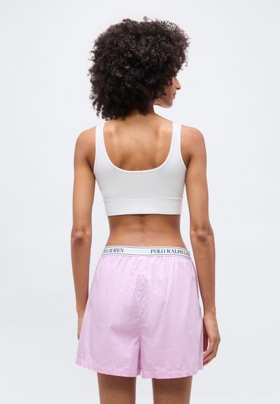 Γυναίκα με σγουρά μαλλιά που φοράει ένα λευκό crop τοπ και ροζ-λευκά ριγέ σορτς Polo Ralph Lauren, στέκεται γυρισμένη προς τα πίσω.