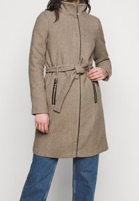 Cappotto marrone, testurizzato, con colletto alto e cintura. Presenta tasche nere con zip e un design aderente. Indossato sopra pantaloni di jeans blu.