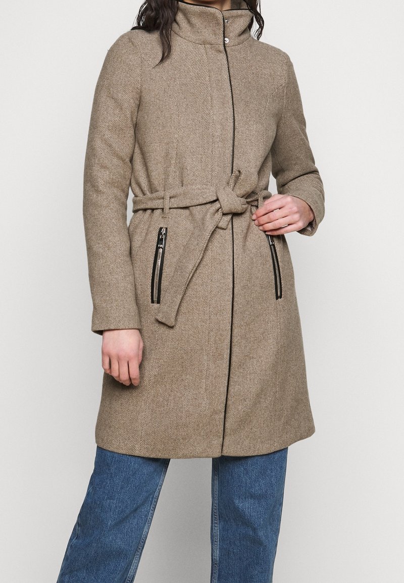 Cappotto marrone, testurizzato, con colletto alto e cintura. Presenta tasche nere con zip e un design aderente. Indossato sopra pantaloni di jeans blu.