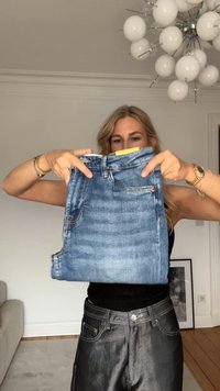 Blå denimjeans hålls upp, med fem fickor, kontrasterande sömmar och en gul etikett vid midjebandet. Texturen är jämn och av medelvikt.