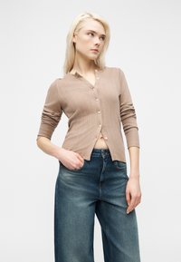 Ξανθιά γυναίκα που φοράει μπεζ ζακέτα με κουμπιά και μπλε τζιν παντελόνι με φαρδιά μπατζάκια, με το χέρι στην τσέπη, κοιτάζοντας προς τα πλάγια σε απλό φόντο.