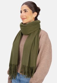 Écharpe vert olive en matière douce, présentant un design simple avec des bords frangés. Portée lâchement autour du cou.