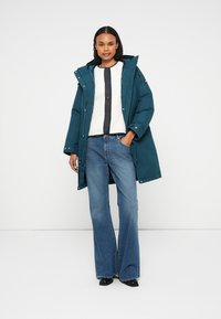 Parka isolada em verde-água com capuz, apresentando botões de pressão e bolsos laterais. Usada sobre um suéter branco com um detalhe de botão escuro e jeans flare azuis.