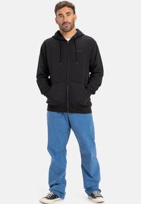 Sweatshirt preto com fecho, cordões, bolsos frontais e um logótipo discreto; combinado com calças azul claro e sapatilhas pretas. Design clean.