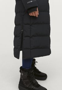 Sort polstret lang jakke med lynlås-lommer i siden; parret med sorte støvler foret med pels, der har snørebånd og et mønstret design. Blå denim synlig.